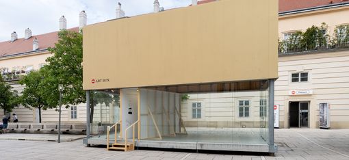 Glass container with beige-clad upper section and wooden stairs in front of a historic building on a paved square