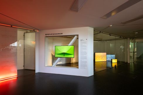 Moderner Ausstellungsraum mit weißer Wand, auf der ein Bildschirm mit grünem Bild hängt, und mehreren beleuchteten rechteckigen Objekten im Hintergrund.