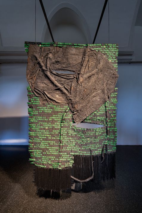 Großes, hängendes Kunstwerk aus grobem, braunem Stoff mit grünen Zahlen und schwarzen Fransen in einem Raum mit gewölbter Decke