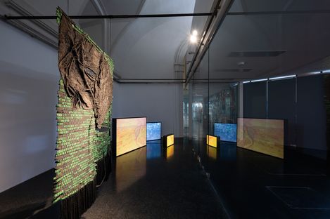 Installationsraum mit einem großen, vertikal hängenden Textilkunstwerk und mehreren leuchtenden rechteckigen Bildschirmen auf dem Boden, die an einer Glaswand reflektiert werden