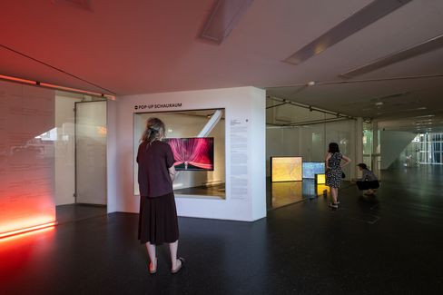 Zwei Personen betrachten in einem modernen Ausstellungsraum digitale Kunstwerke auf Bildschirmen, eine weitere Person kniet vor einem Bildschirm im Hintergrund