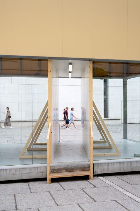 Durch eine Glasfront mit Durchgang sind drei Personen zu sehen, die auf einem gepflasterten Platz an einer weißen Wand vorbeigehen.