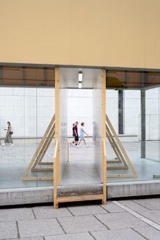 Durch eine Glasfront mit Durchgang sind drei Personen zu sehen, die auf einem gepflasterten Platz an einer weißen Wand vorbeigehen.