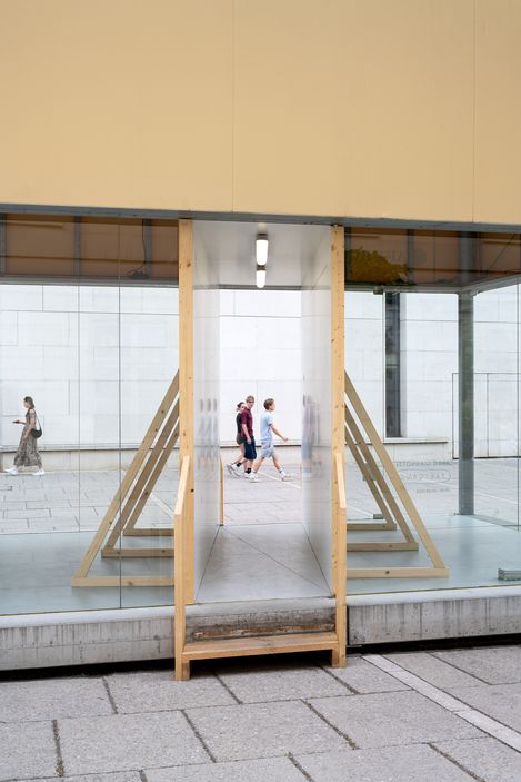 Durch eine Glasfront mit Durchgang sind drei Personen zu sehen, die auf einem gepflasterten Platz an einer weißen Wand vorbeigehen.