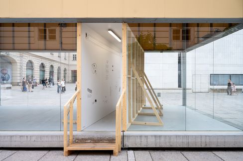 Glasgebäude mit Durchgang und Treppe, im Hintergrund Menschen auf einem Platz vor einem historischen Gebäude