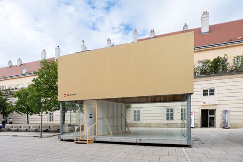 Glascontainer mit beige verkleideter Oberseite und Holztreppe vor einem historischen Gebäude auf einem gepflasterten Platz