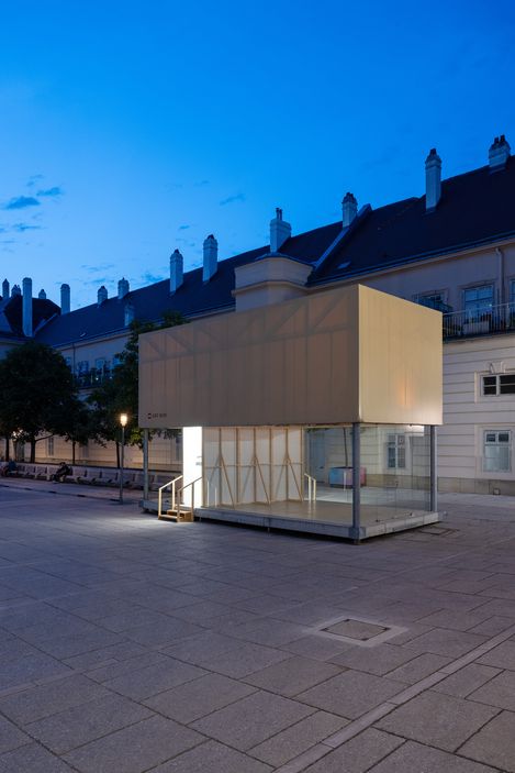 Kubischer Pavillon mit verglaster Unterseite und geschlossener Oberseite auf gepflastertem Platz vor historischen Gebäuden bei Dämmerung