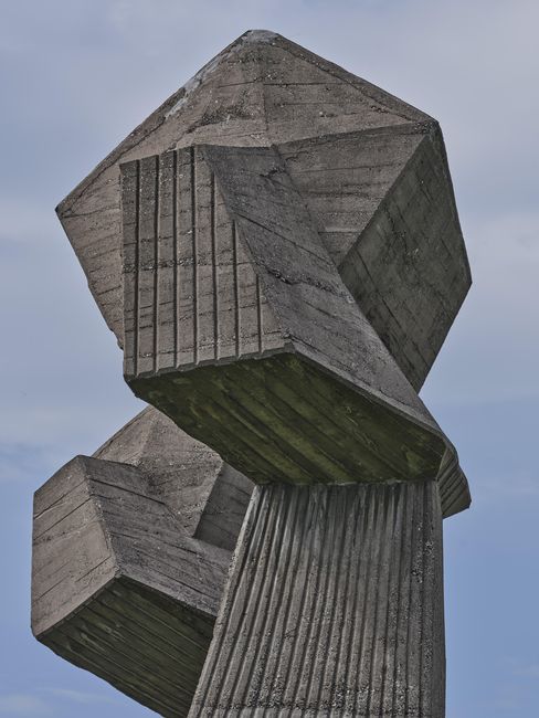 Betonskulptur mit zwei kubischen Formen auf einem gerillten, zylindrischen Sockel vor blauem Himmel.