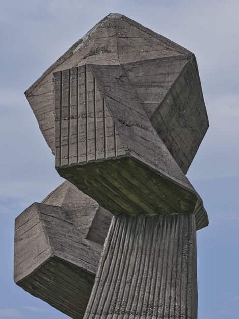 Betonskulptur mit zwei kubischen Formen auf einem gerillten, zylindrischen Sockel vor blauem Himmel.