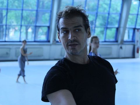 Man in a dark t-shirt in a large room with big windows, blurred people in the background