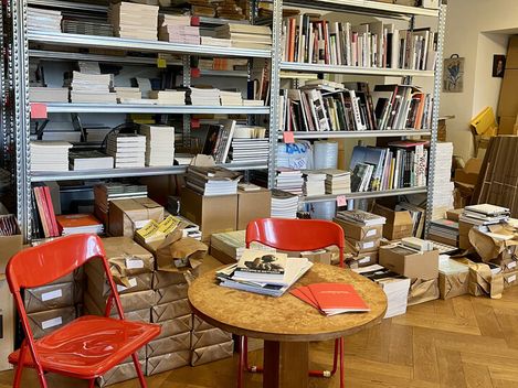 Kleiner runder Holztisch mit Büchern und Heften, umgeben von zwei roten Stühlen, vor Regalen mit gestapelten Büchern und braunem Packpapier.