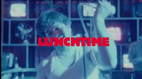 Eine Person singt auf einer Bühne in ein Mikrofon, blau beleuchtet. In roter Schrift steht „LUNCHTIME“ über dem Bild.