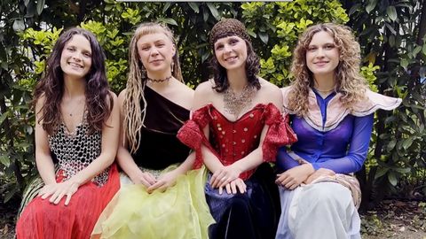 Vier Personen sitzen nebeneinander im Freien vor grünen Sträuchern und tragen auffällige, farbenfrohe Outfits.
