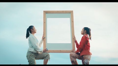 Zwei Frauen knien sich gegenüber und halten gemeinsam einen großen, leeren Holzrahmen vor einem hellen Himmel