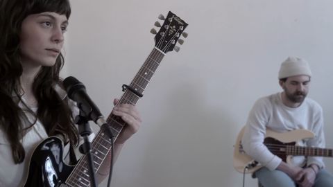 Frau mit dunklen Haaren hält eine E-Gitarre und steht vor einem Mikrofon, im Hintergrund sitzt ein Mann mit weißer Mütze und hält eine E-Gitarre