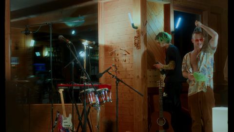 Zwei Männer in einem Raum mit Holzwänden, einer mit grünen Haaren schaut auf ein Handy, der andere trägt eine bunte Hemdjacke und hält eine Schüssel, davor stehen Mikrofone, Gitarren und ein Schlagzeug.