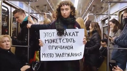 Frau mit lockigem Haar und Schal hält in einer vollen U-Bahn einen handgeschriebenen Zettel mit russischem Text.