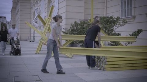 Zwei Personen tragen gelbe Stahlträger vor einem hellen Gebäude auf einem gepflasterten Platz.