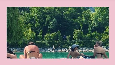 Drei Frauen in Badebekleidung sitzen am Rand eines Gewässers mit grünem Wasser, im Hintergrund Bäume und Steine.
