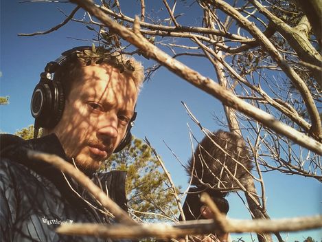 Person wearing headphones holding microphone with windscreen near bare tree branch against clear sky.