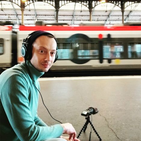 Person mit Kopfhörern und grünem Pullover sitzt auf Bahnsteig vor einem fahrenden Zug.