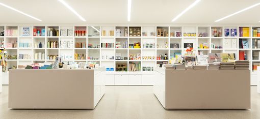 Symmetrical wide view of a modern museum shop with white furniture, shelving and a diverse product range.