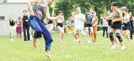 Frau mit langem blondem Haar tanzt barfuß auf einer Wiese vor einer Gruppe von Menschen, die ihr folgen oder zuschauen