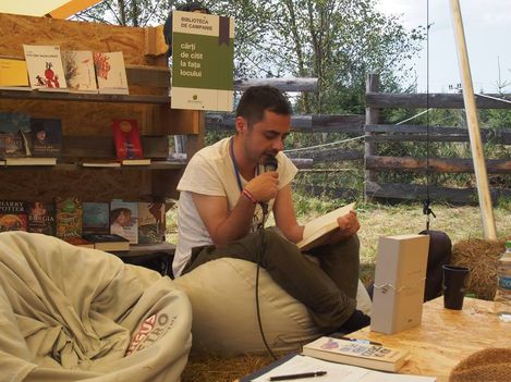 Person sitting on a beanbag inside a tent reading from a book surrounded by shelves with books and a sign reading "BIBLIOTECA DE CAMPANIE cărți de citit la fața locului".