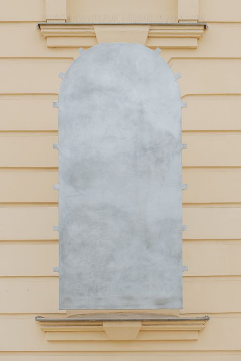 Closed, light grey, arched metal art installation in a window recess on a light yellow façade with horizontal lines