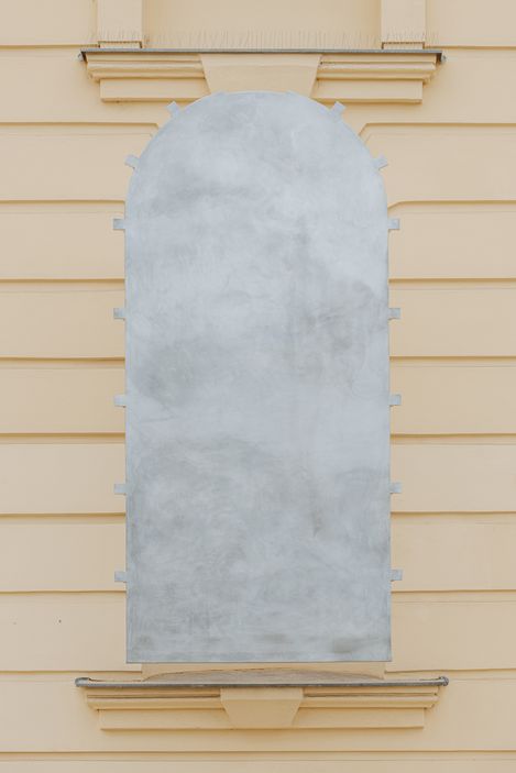Closed, light grey, arched metal art installation in a window recess on a light yellow façade with horizontal lines