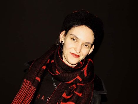 Person with dark hair wearing a red-and-black scarf and dark clothing against a black background.