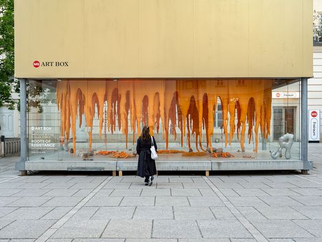 A person in black stands facing a glass art box displaying hanging orange sculptures and abstract shapes titled "Roots of Resistance.