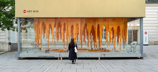 A person in black stands facing a glass art box displaying hanging orange sculptures and abstract shapes titled "Roots of Resistance.