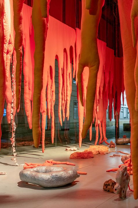 Long orange organic-shaped sculptures hang from the ceiling in an exhibition space with additional sculptural objects on the floor.