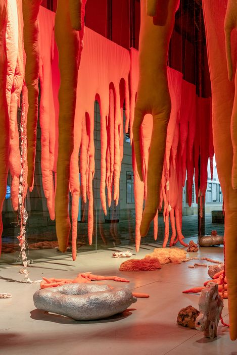 Long orange organic-shaped sculptures hang from the ceiling in an exhibition space with additional sculptural objects on the floor.