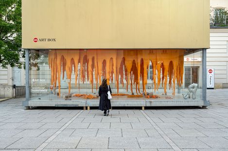 A person in black stands facing a glass art box displaying hanging orange sculptures and abstract shapes titled "Roots of Resistance.