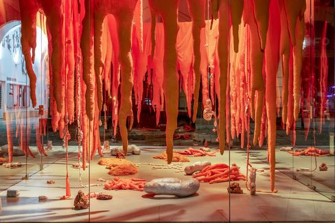 An artistic exhibit features vibrant orange fabric hanging like organic forms, resembling tentacles. The installation includes scattered objects, under glass with a dramatic lighting.