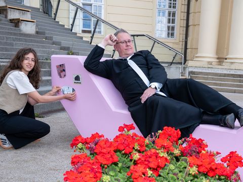 A young woman attaches a sticker to the pink Enzi at Klosterneuburg Abbey. Beside her, a clergyman reclines on the seating furniture. Red geraniums bloom in the foreground, while steps leading to the Baroque abbey complex can be seen in the background.