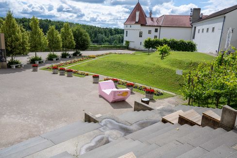 View from a staircase across the grounds of Klosterneuburg Abbey. The pink Enzi stands among flower beds and landscaped lawns, surrounded by historic buildings and a row of trees at the edge of the site.