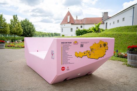 The pink Enzi at Klosterneuburg Abbey featuring the “Enzi on Tour” graphic and a map of Austria showing the stops on its journey. Lawns, flower beds and the historic abbey buildings can be seen in the background.