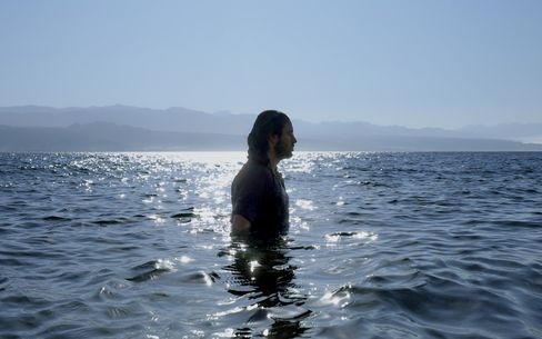 Person standing waist-deep in a lake or sea with mountains in the background and sunlight reflecting on the water surface.