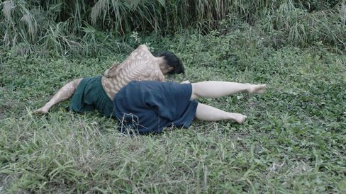 A person lies on a grassy meadow in a twisted, ground-level position that suggests movement or performance. Their upper body is painted with light, plant-like patterns, while the background is filled with dense greenery. The scene feels calm yet physically intense.