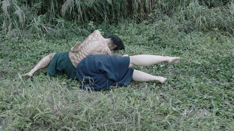 A person lies on a grassy meadow in a twisted, ground-level position that suggests movement or performance. Their upper body is painted with light, plant-like patterns, while the background is filled with dense greenery. The scene feels calm yet physically intense.