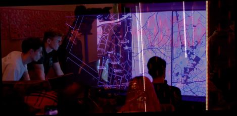 Several people sit in front of a large vertical projection of city maps showing streets and rivers in a dark room.