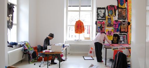 Zwei Personen arbeiten in einem hellen Atelier mit bunten T-Shirts und Drucken an der Wand.