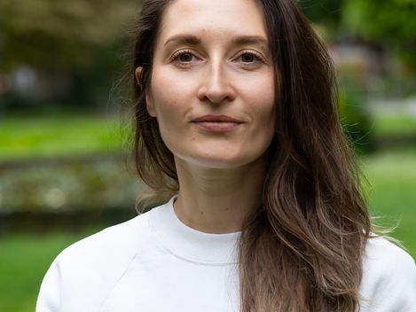 Person with long brown hair wearing a white sweater standing in front of a blurred background with trees and buildings.