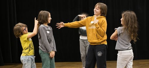 Auf dem Bild sind 5 Kinder zu sehen, die offensichtlich gerade eine Szene spielen: Eines hat den Arm ausgestreckt, ein anders die Arme verschränkt, ein drittes verbirgt sich hinter einem vierten und ein fünftes im Hintergrund hält Ausschau nach den anderen.