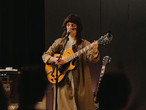 Person mit langem Mantel spielt E-Gitarre auf dunkler Bühne.