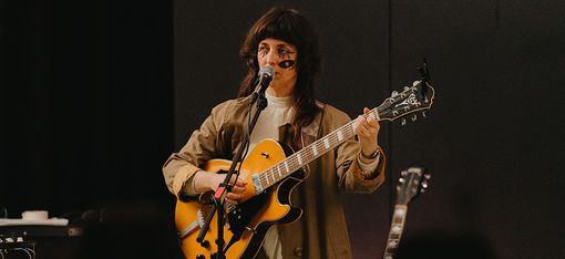 Person mit langem Mantel spielt E-Gitarre auf dunkler Bühne.