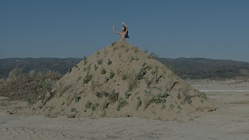 A person stands on a large sand mound with sparse vegetation raising one arm against a background of hills and blue sky.